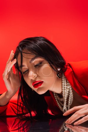 Stilvolle brünette Frau in Perlenkette berühren reflektierende Oberfläche isoliert auf rot 
