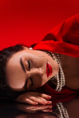 Photo for Young woman in red jacket and pearl necklace lying on reflective surface on red background - Royalty Free Image