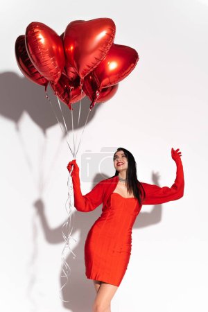 Cheerful brunette woman in red dress looking at heart shaped balloons on white background with shadow 