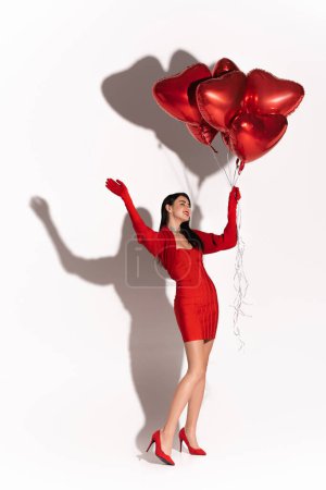 Cheerful woman in heels and dress holding red heart shaped balloons and waving hand on white background with shadow 
