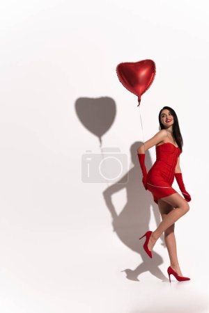 Photo for Sexy brunette woman in heels and red gloves holding heart shaped balloon on white background with shadow - Royalty Free Image