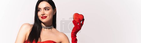 Foto de Mujer morena de moda con labios rojos sosteniendo regalo en forma de corazón sobre fondo gris, pancarta - Imagen libre de derechos
