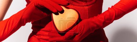 Vue partielle de la femme en gants rouges tenant boîte cadeau en forme de coeur sur fond gris, bannière 