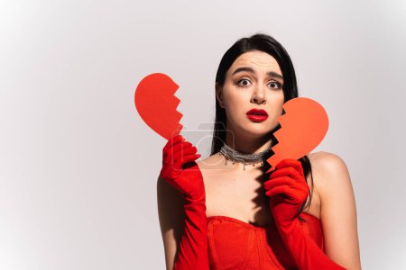 Téléchargez les photos : Femme inquiète en gants rouges tenant le cœur en papier cassé isolé sur gris - en image libre de droit