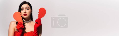 Téléchargez les photos : Femme inquiète dans des gants rouges tenant le coeur brisé de papier isolé sur gris, bannière - en image libre de droit