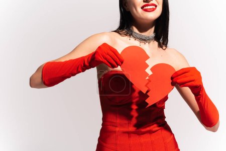 Téléchargez les photos : Vue recadrée d'une femme souriante avec des lèvres rouges tenant du papier cœur brisé isolé sur gris - en image libre de droit
