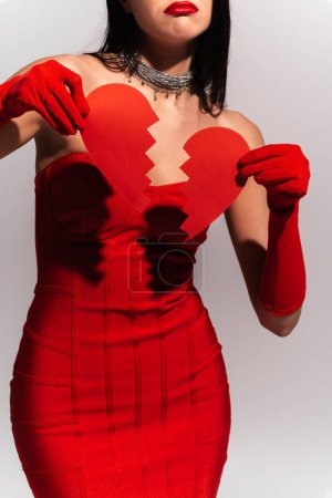 Téléchargez les photos : Vue recadrée de femme élégante bouleversée en robe rouge tenant coeur en papier cassé isolé sur gris - en image libre de droit
