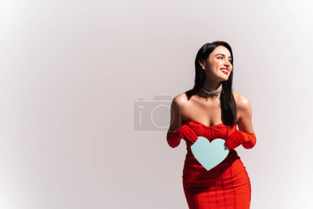 Cheerful woman in red dress with naked shoulders holding paper heart isolated on grey 