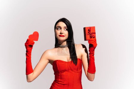 Photo for Pensive woman in red dress holding paper heart and gift isolated on grey - Royalty Free Image