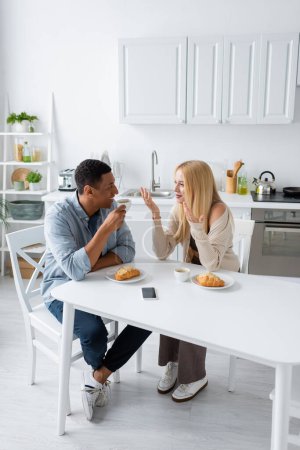 happy interracial couple talking near coffee cups and croissants while having breakfast in kitchen