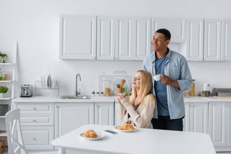 Photo for Cheerful multiethnic couple with coffee cups looking away near delicious croissants in kitchen - Royalty Free Image