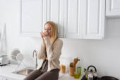 blonde woman with closed eyes sitting on kitchen worktop and enjoying flavor of morning coffee puzzle #625474400