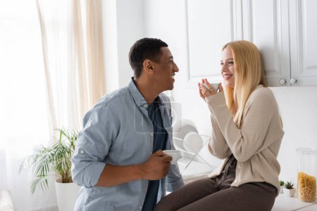 smiling blonde woman with coffee cup sitting on kitchen worktop near african american boyfriend