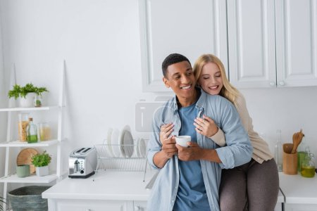 joyful blonde woman with closed eyes sitting on kitchen worktop and hugging african american boyfriend standing with coffee cup