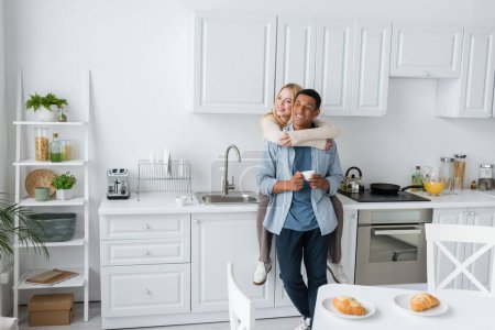 happy woman sitting on worktop in modern kitchen and embracing african american boyfriend with coffee cup