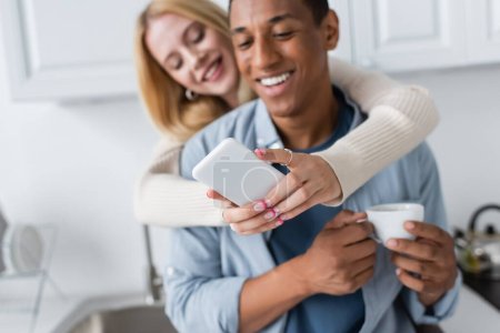 foyer sélectif du smartphone dans les mains de la femme floue près de l'homme afro-américain avec tasse de café
