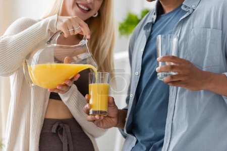 partial view of smiling woman pouring orange juice near african american boyfriend with glasses 