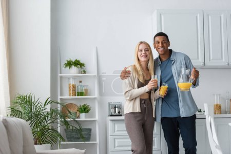 joyful interracial couple holding orange juice and looking at camera in modern kitchen