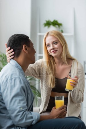 Téléchargez les photos : Heureux jeune femme tenant verre de jus d'orange frais et toucher la tête de l'homme afro-américain dans la cuisine - en image libre de droit