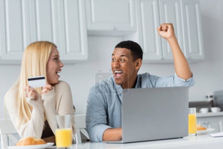 Photo pour Excité homme afro-américain se réjouissant près ordinateur portable et petite amie avec carte de crédit pendant le petit déjeuner dans la cuisine - image libre de droit