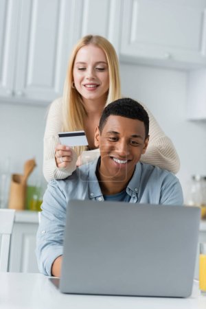 Téléchargez les photos : Femme blonde tenant carte de crédit près de sourire copain afro-américain et ordinateur portable dans la cuisine - en image libre de droit
