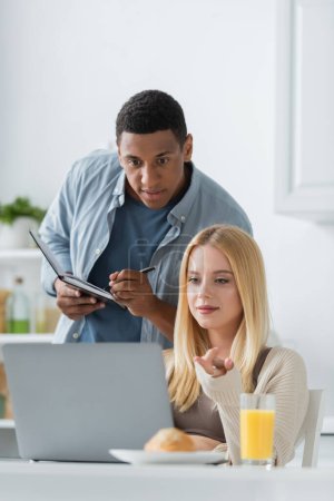 Photo for African american man writing in notebook near positive girlfriend pointing at laptop in kitchen - Royalty Free Image