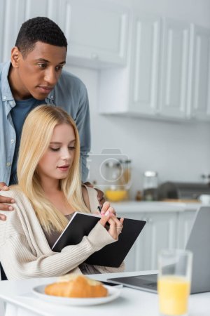 Photo for African american man hugging shoulders of blonde girlfriend writing in notebook near laptop and blurred breakfast - Royalty Free Image
