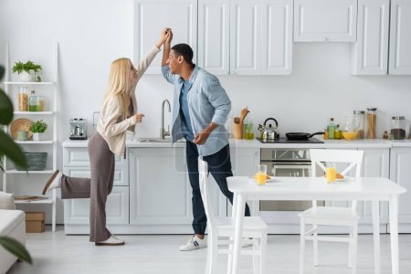 full length of cheerful multiethnic couple holding hands and looking at each other while dancing in kitchen