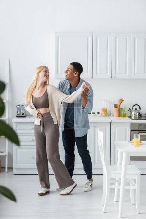 full length of joyful interracial couple smiling at each other while dancing in modern kitchen