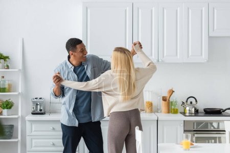 smiling african american man dancing with young blonde girlfriend in kitchen