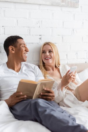 glückliches gemischtrassiges Paar mit Buch und Handy, die einander im Schlafzimmer anschauen