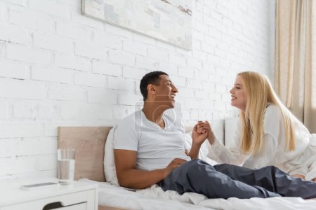 joyful multiethnic couple holding hands and looking at each other on bed