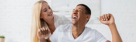Photo pour Excité homme afro-américain avec les yeux fermés riant près souriant femme blonde dans la chambre, bannière - image libre de droit