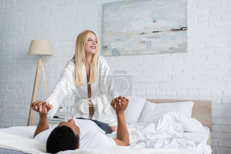 Foto de Mujer feliz en túnica de seda blanca mirando hacia otro lado y tomándose de la mano con el hombre afroamericano acostado en la cama - Imagen libre de derechos