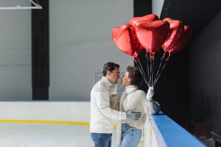 Téléchargez les photos : Vue latérale d'une femme afro-américaine joyeuse embrassant son petit ami et tenant des ballons en forme de cœur sur une patinoire - en image libre de droit