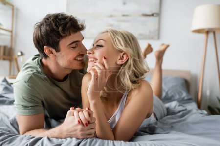 Smiling man in pajama kissing and holding hand of cheerful girlfriend on bed 