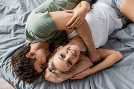 Foto de Vista superior de un hombre alegre abrazando a su novia en pijama mirando la cámara en la cama - Imagen libre de derechos