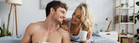 Téléchargez les photos : Femme blonde gaie tenant une tasse de café près de copain torse nu dans la chambre, bannière - en image libre de droit
