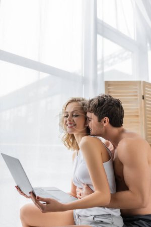 Téléchargez les photos : Jeune homme musclé étreignant petite amie blonde gaie avec ordinateur portable à la maison - en image libre de droit