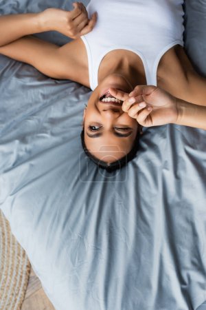 Photo for Top view of cheerful african american woman winking while lying on bed at home - Royalty Free Image