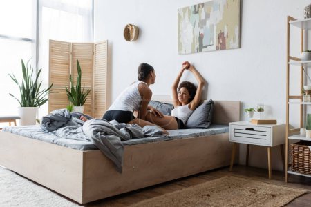 young lesbian african american woman looking at happy girlfriend with raised hands in modern bedroom
