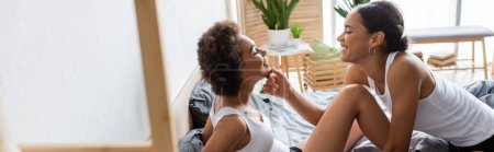 happy lesbian african american woman touching chin of young girlfriend in modern bedroom, banner