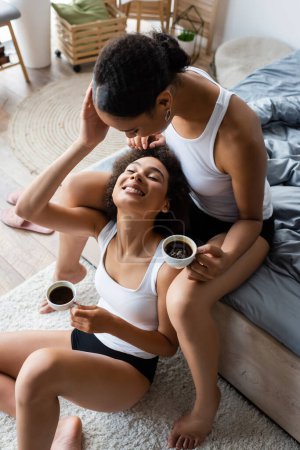 Draufsicht auf fröhliche lesbische afrikanisch-amerikanische Frauen, die sich im Schlafzimmer umarmen und Tassen Kaffee halten 