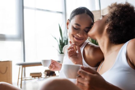 lesbische Afroamerikanerin küsst lächelnde Freundin mit Tasse Kaffee auf die Wange 