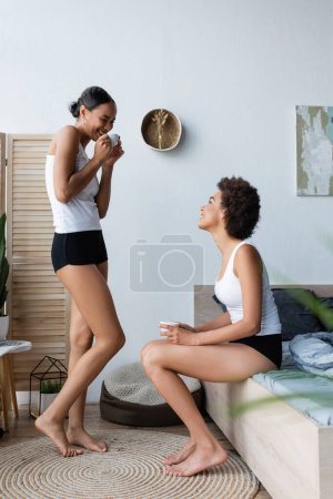 Téléchargez les photos : Africaine américaine lesbienne femme boire café tandis que debout près de petite amie dans chambre - en image libre de droit
