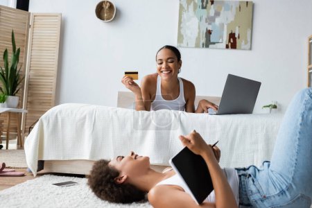 junge afrikanisch-amerikanische Frau liegt auf Teppich und schaut glückliche Freundin mit Kreditkarte an, während sie Laptop benutzt 