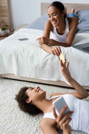 young african american woman lying on carpet and giving credit card to girlfriend 