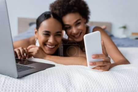 sonriente afroamericana lesbiana mujer mostrando smartphone a novia cerca de portátil en la cama