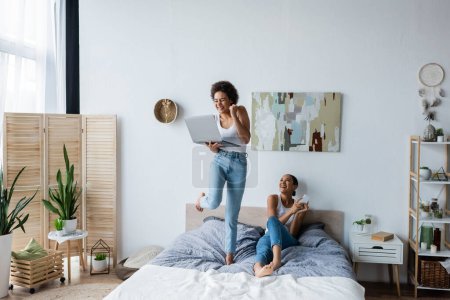 excited african american lesbian woman holding laptop while standing on bed near smiling girlfriend  