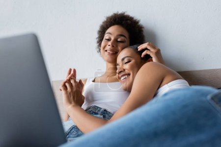 happy african american lesbian couple watching movie on laptop and holding hands in bedroom